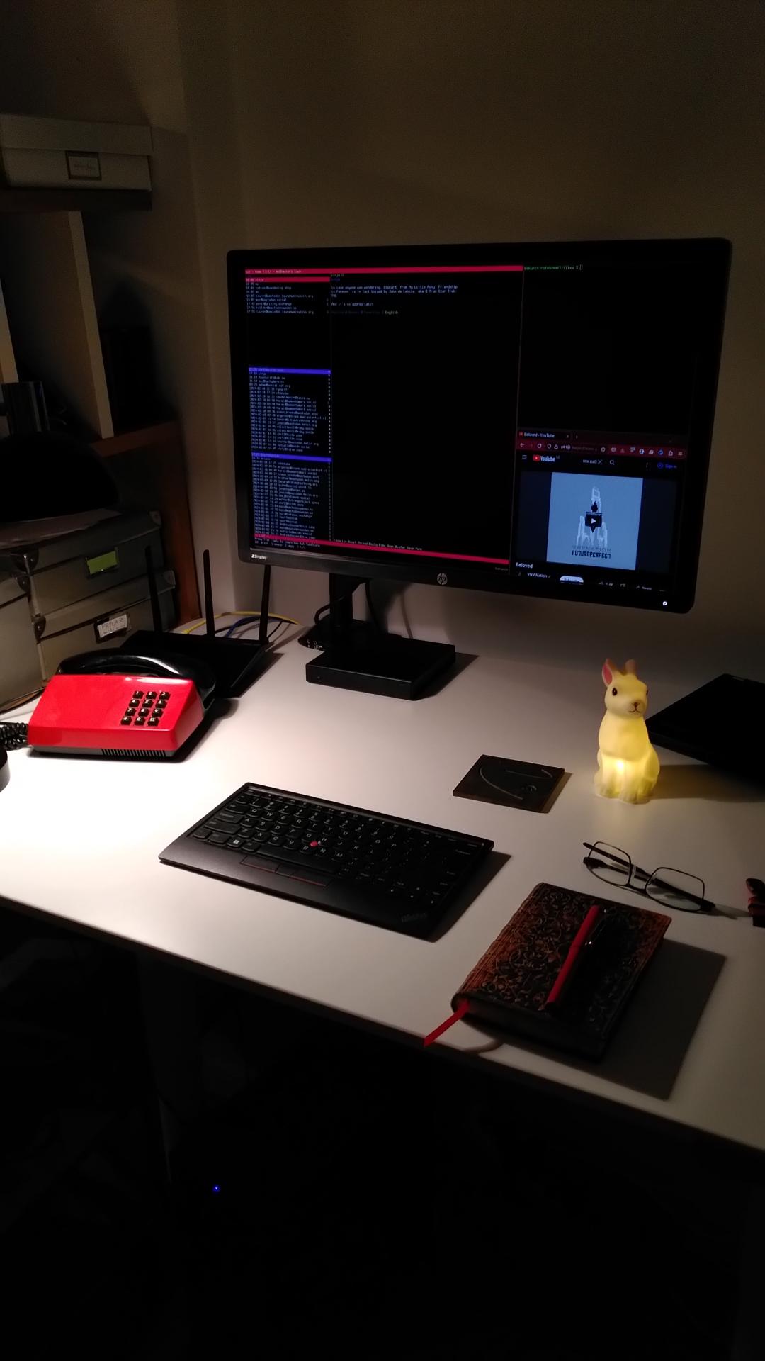 Small white desk with a red/black classical touchtone phone, a 24" monitor on an arm, a small lit bunny children's light, a fancy notebook with a pen, a pair of glasses