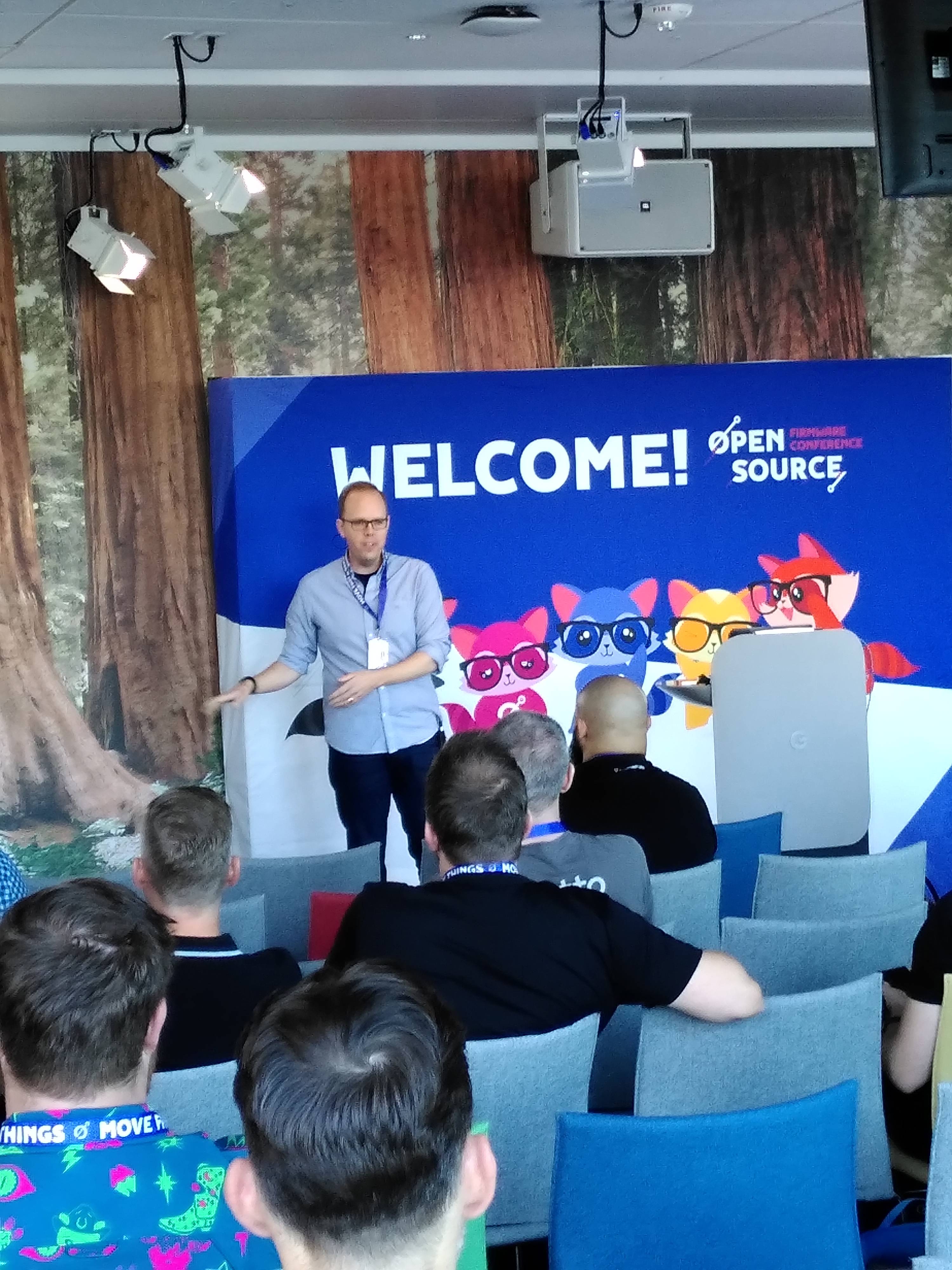 White guy with blue shirt and black trousers on the podium in the main OSFC room gesticulating while talking. Backdrop says "Welcome! And something about "Open Source".