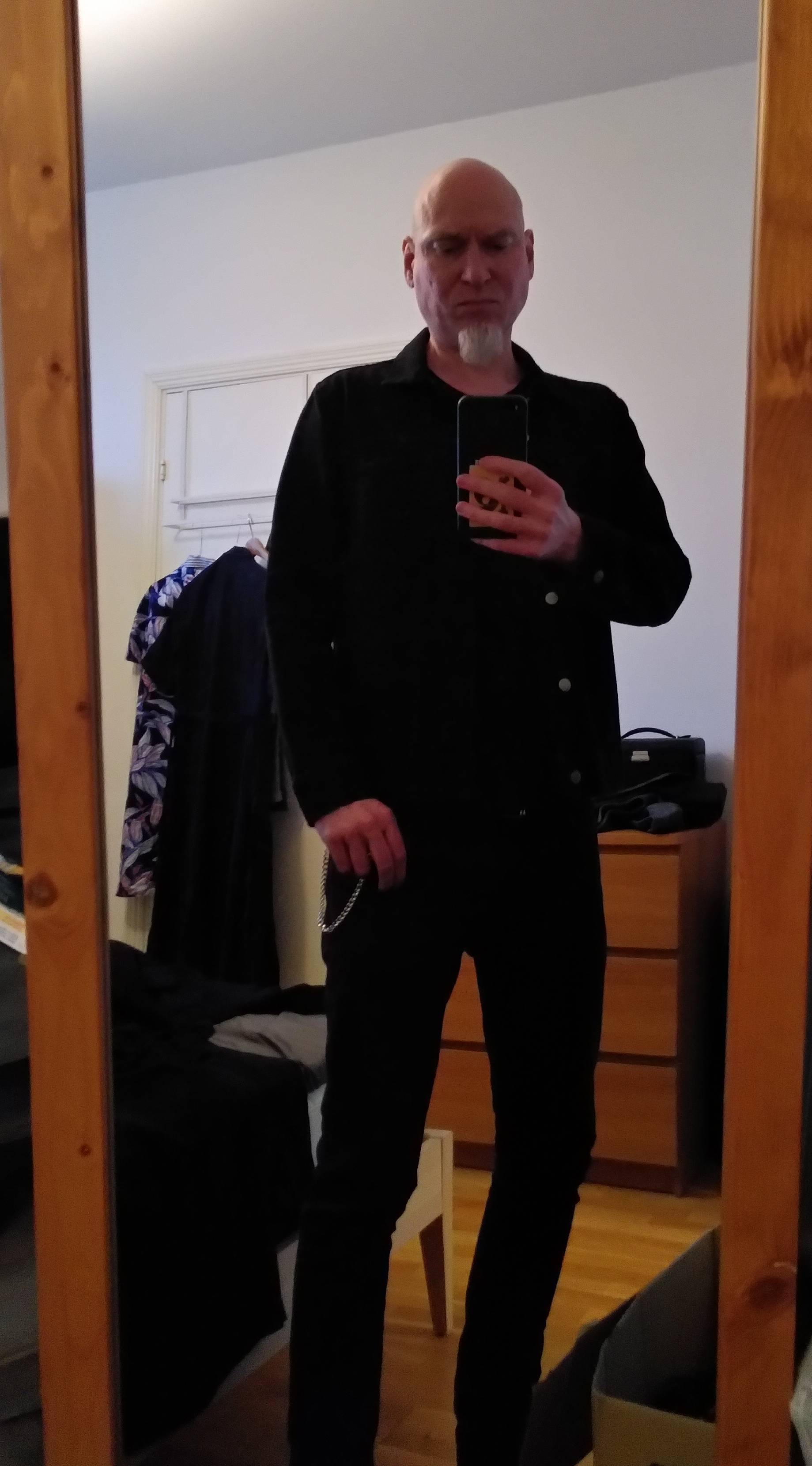 Mirror image of a white, bald man with a white goatee in black denim jacket, black t-shirt and black denim trousers with a key chain. Eyes looking down, so no eye contact.