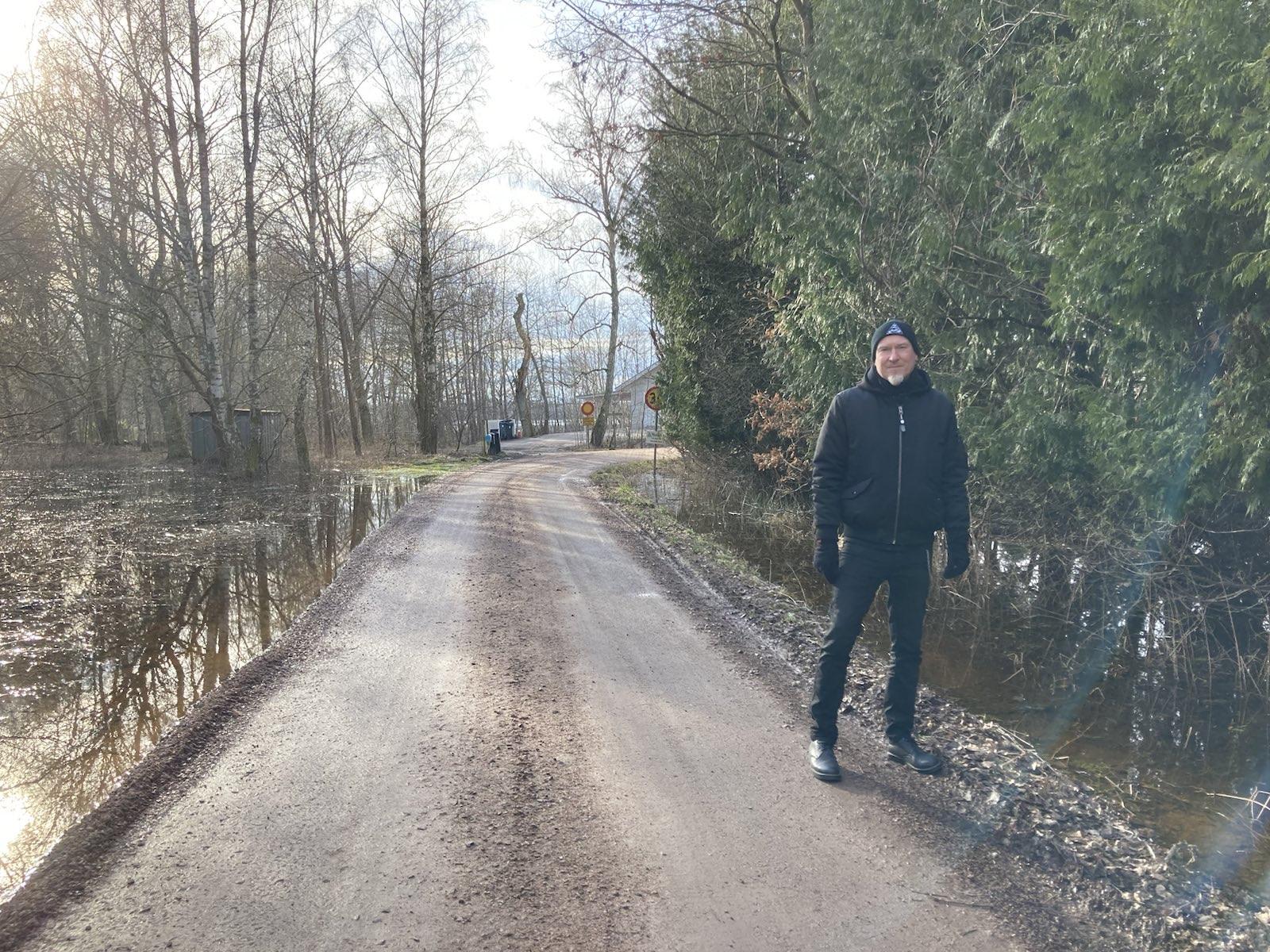 Me, a pale male in black clothing, standing on a gravel road with lots of water in the forest on both sides.
