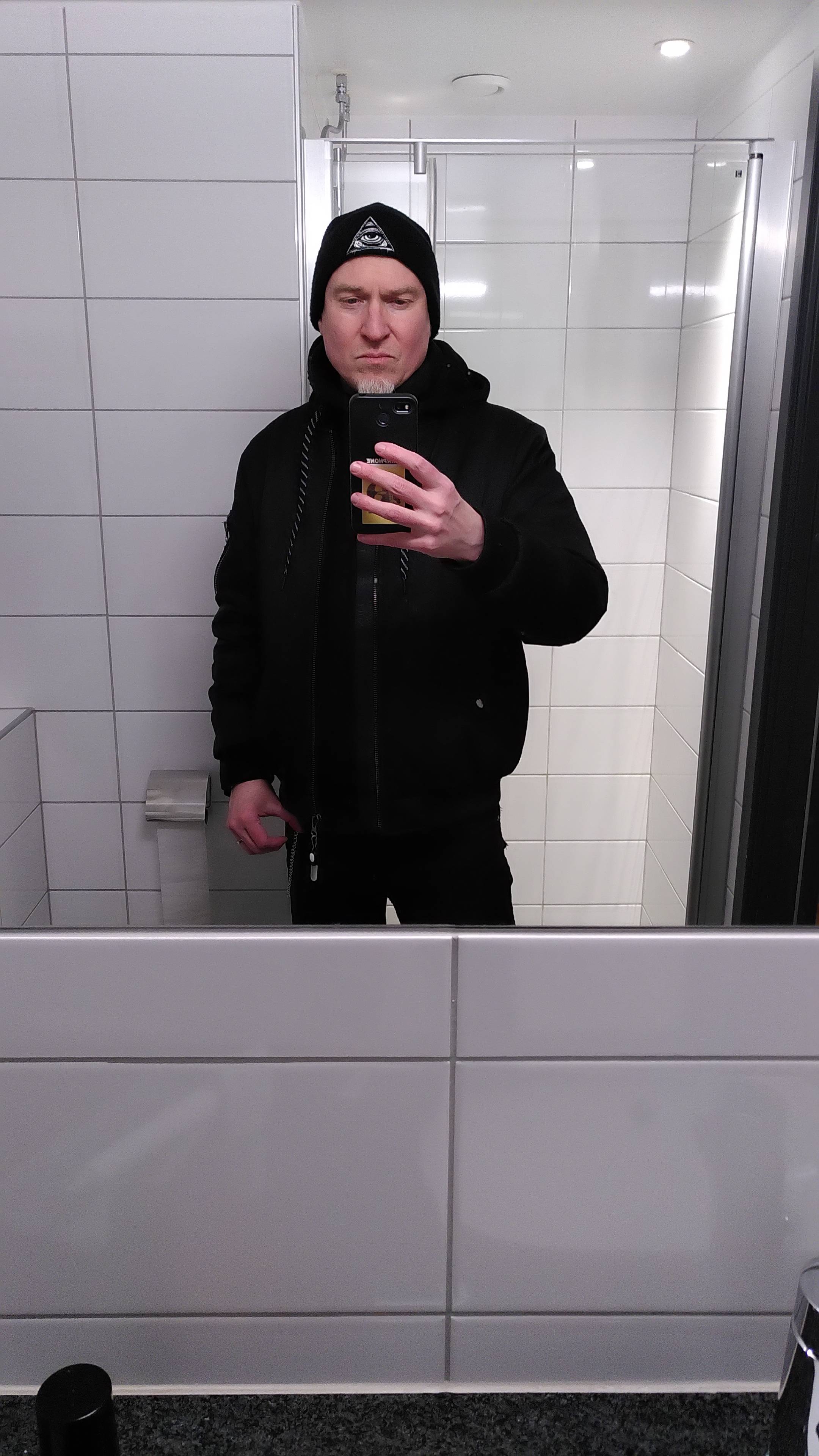Me, a pale guy with a white goatee, dressed in black with a black bomber and black beanie, as seen in a bathroom mirror.