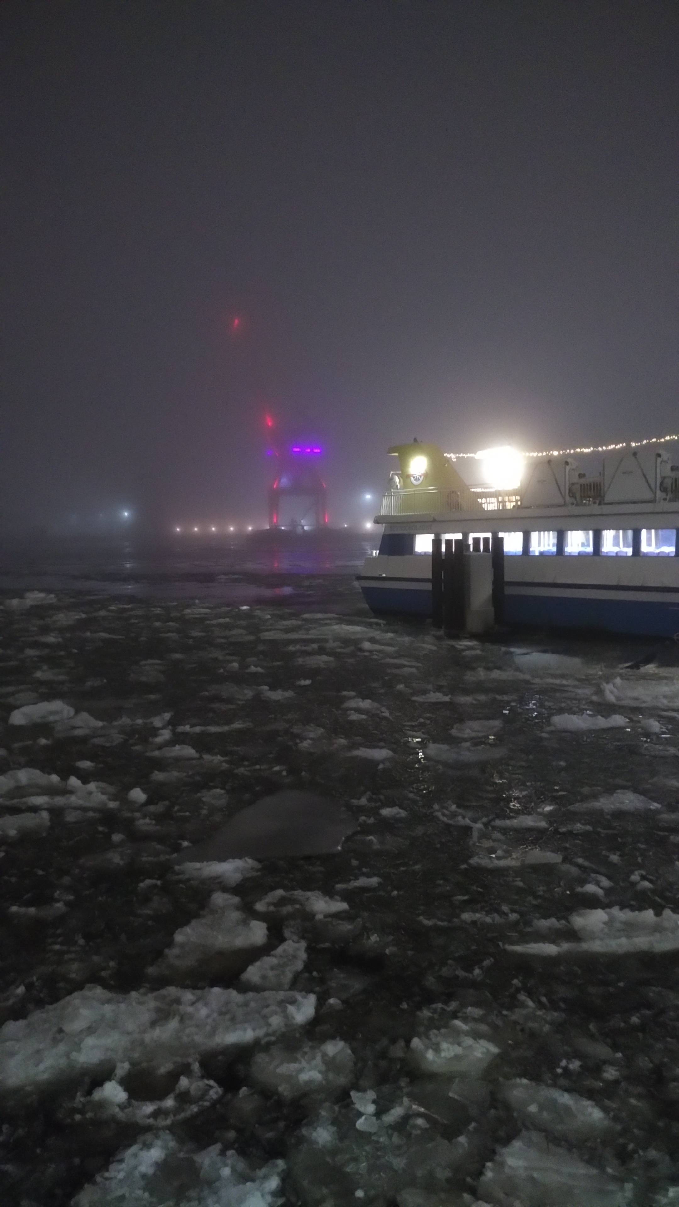 An icy river with a ferry just arriving. In the background you can almost see a crane decorated with purple lights through the fog.