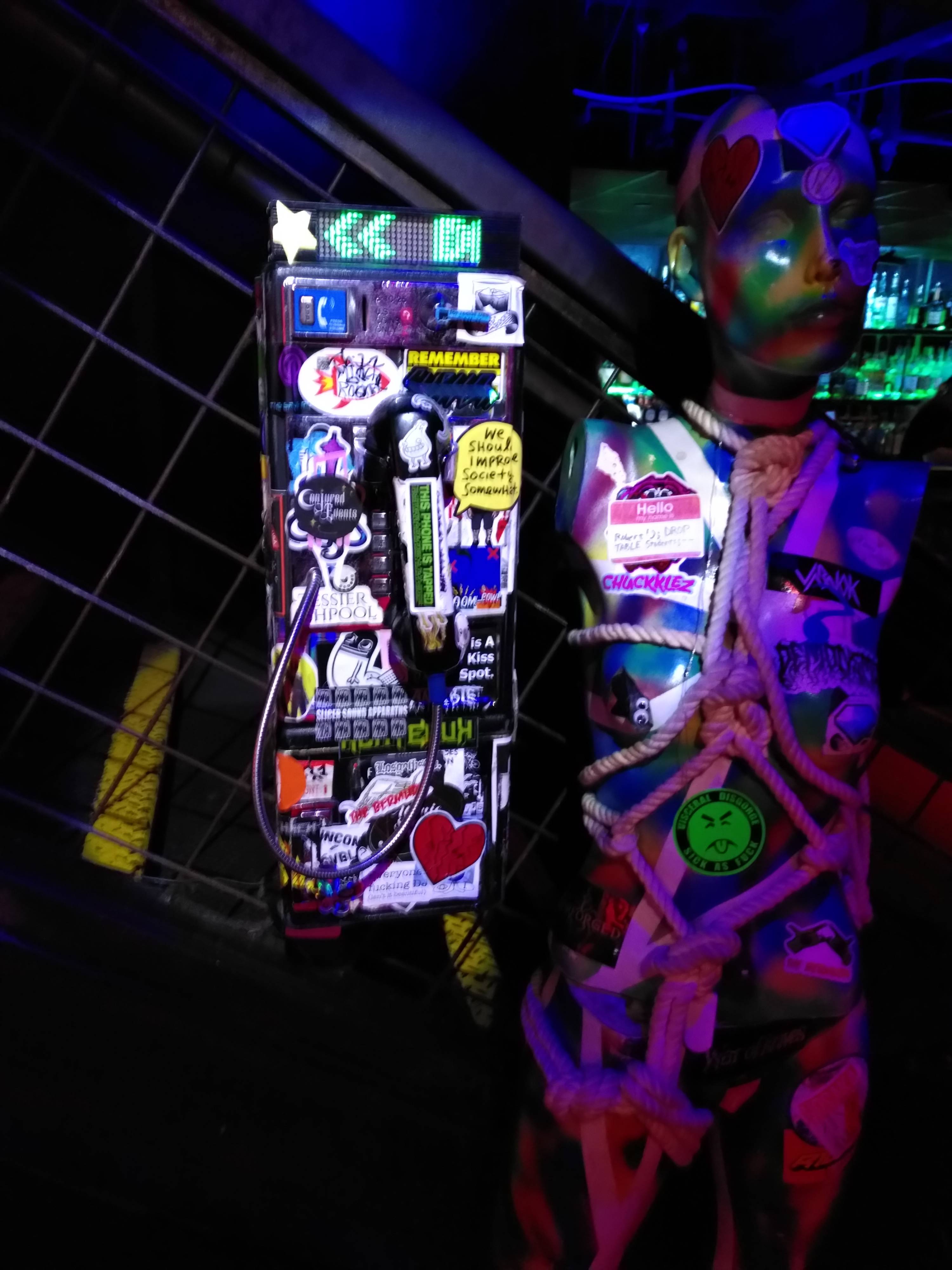 An american payphone covered in stickers in neon light. Beside it is a painted mannequin with some shibari going on.