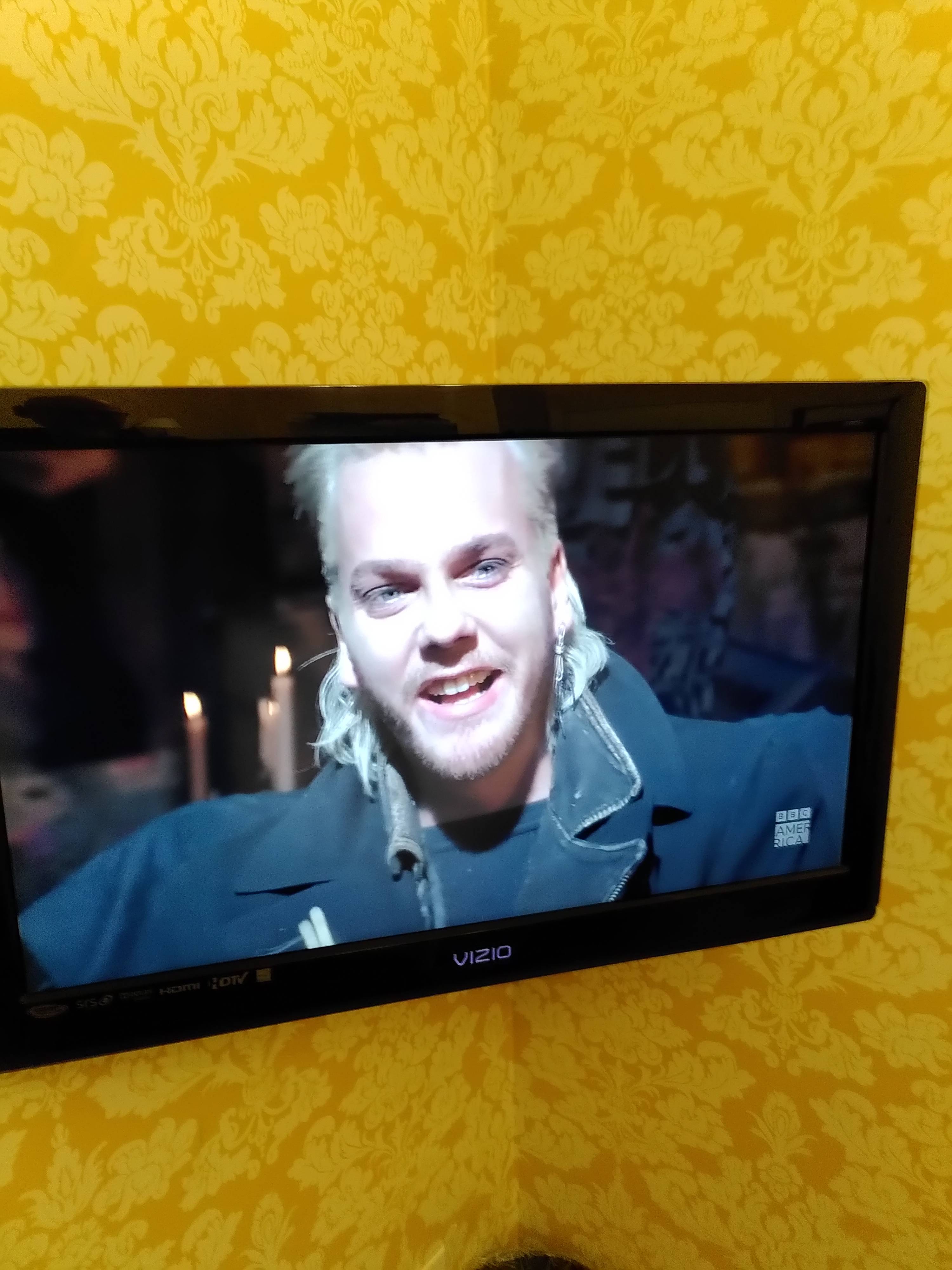A slightly bearded white male in black clothes and a mullet smiling on the TV. Very yellow wall paper behind the TV.