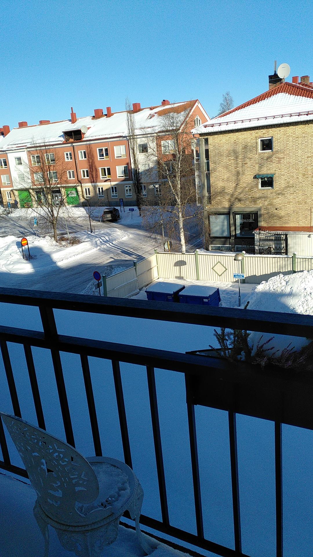 Parts of a balcony, snowy back yard, and a few multi-story houses with snow on the roofs.