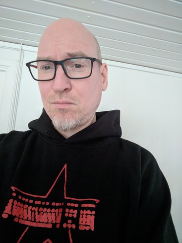 Photo of MC, a pale male with a white beard and red/black glasses wearing a black hoodie with Die Sterntastatur in red"