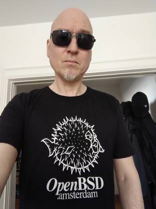 Photo of MC, a pale male with dark sunglasses, wearing a black t-shirt saying "OpenBSD.amsterdam"