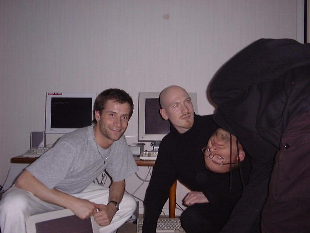 Photo of three men in front of screens: to the left in white trousers, grey shirt, brown hair, to the right: two dressed in black with shaved heads, one with glasses bending over and looking at the camera upside down.