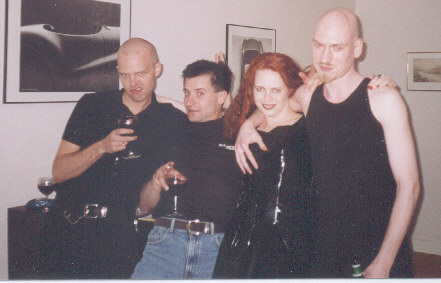 Photo of three men and a woman standing next to each other, most holding some form of alcoholic beverage, all smiling in a way. All are dressed mostly in black. Two of the men have shaved heads, including me, one has black hair. The woman has long red hair and is dressed in a black PVC dress.