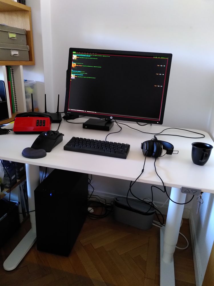 A white desk with a red phone, a black trackball, black small keyboard with blank keycaps, a 24" monitor showing a fullscreen Emacs, headphones. Under the table is a black tower computer standing on a wooden floor.