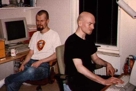 Two pale dudes, one with very short hair and beard in white t-shirt and blue jeas, one with cleanshaven head in short beard dressed in black, both in front of screens