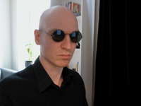 Picture of MC, a cleanshaved, bald man with round, dark sunglasses