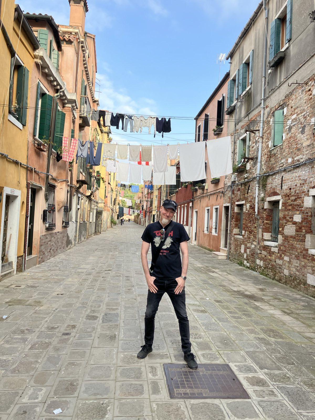 Pale, bald man with white goatee dressed in black pair of jeans and
black band t-shirt, huge grin, standing on a mostly empty very sun-lit
street in Venice, surrounded by old houses, clothing lines above him
and a very blue sky.