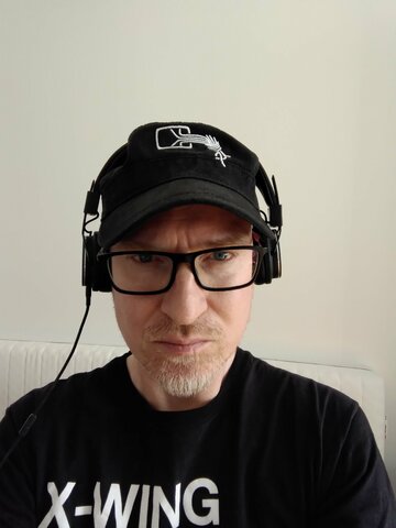 Picture of MC in a military-style black cap with the CCC logo, large, nerdy glasses and a black X-Wing fighter t-shirt staring into the camera