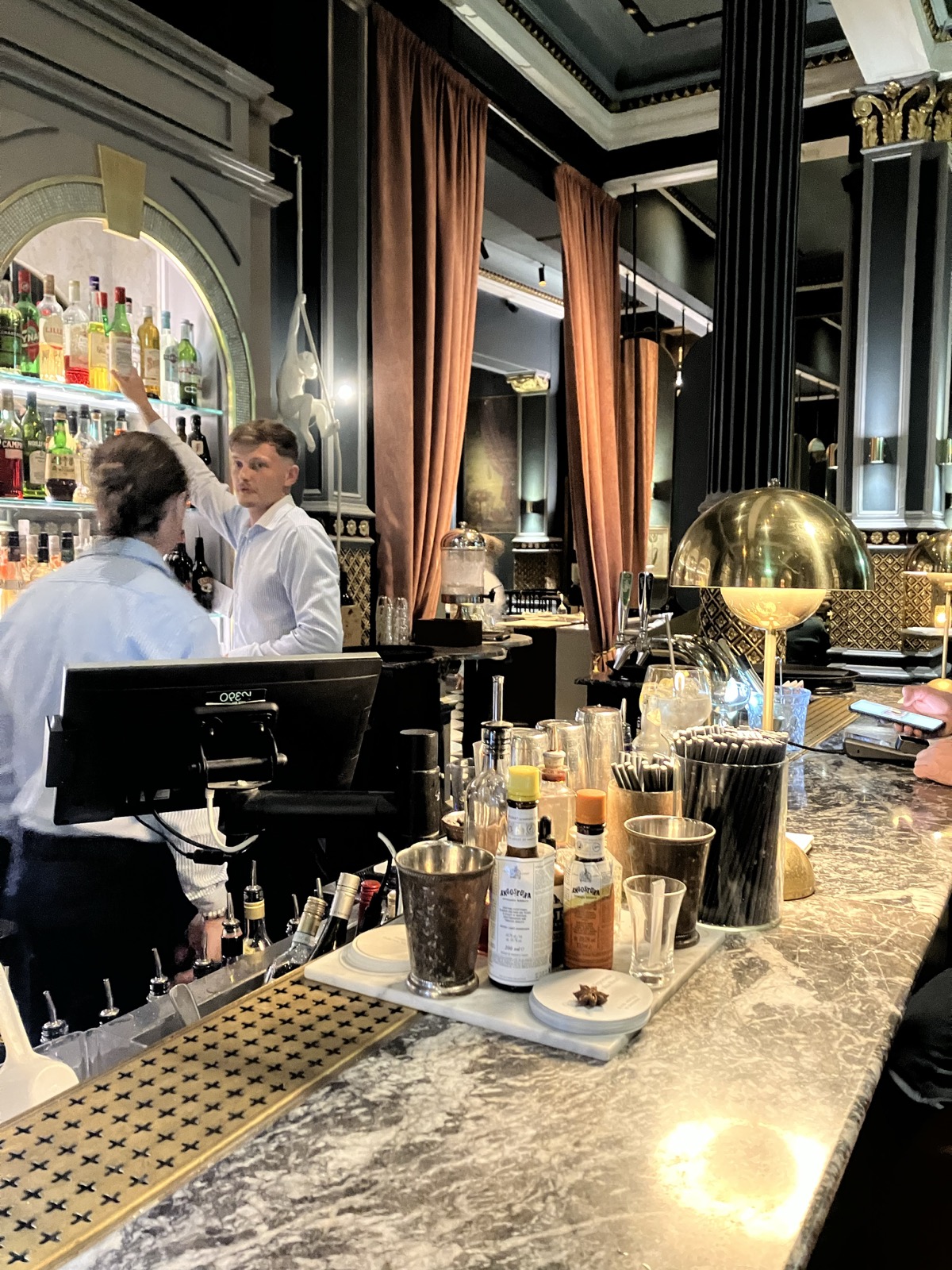 A marble bar disk with a view into the restaurant, painted pillars holding up the roof. Behind the bar, two bartenders in white discussing something. One of them stretching for a bottle.