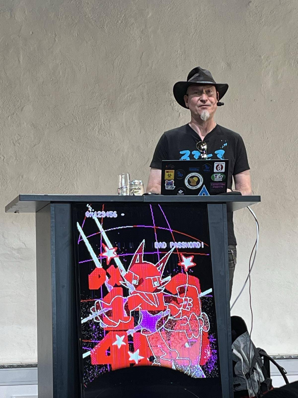 A very pale man dressed in black with a black cowboy hat and a white goatee standing on a stage with a laptop in front of him covered in stickers.