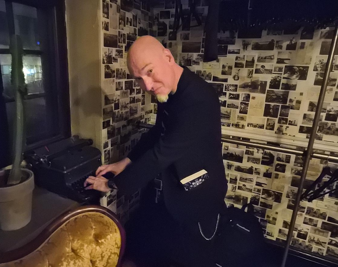 A bald, very pale man with a white goatee dressed completely in black, with a novel in his suit jacket pockets, leans over a mechanical typewriter, looking back the viewer since someone called for him.