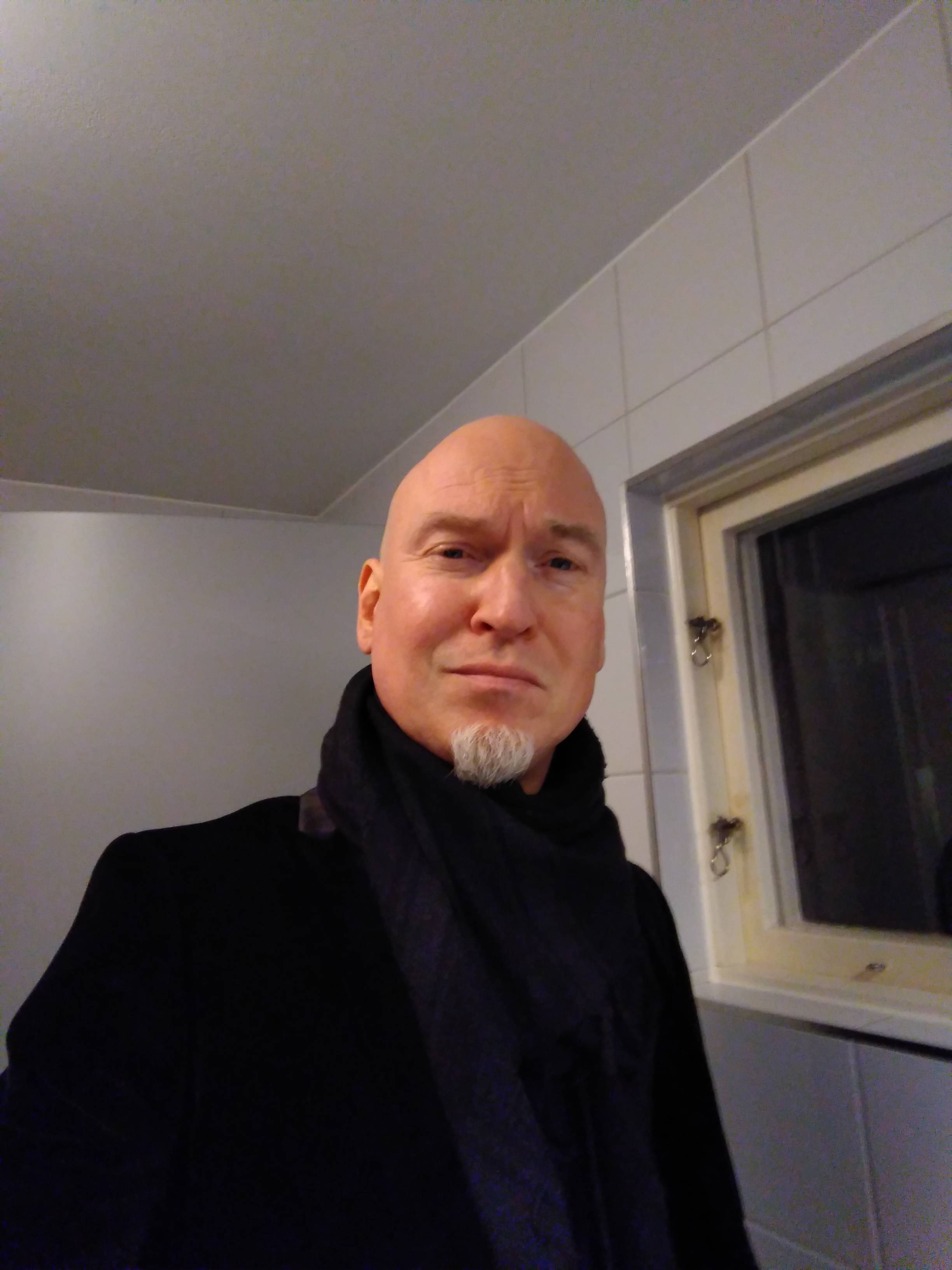 A pale man with shaved head and white goatee in a velvet coat and scarf. Slightly smiling.
