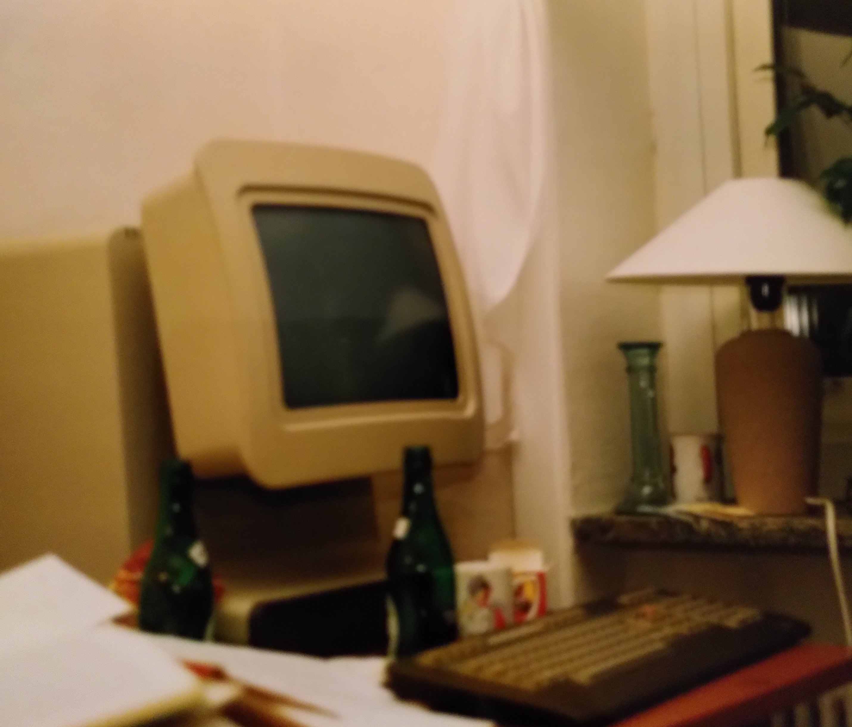 Large beige horisontal stand with a swivel monitor fastened to it standing on a messy desk, a keyboard in front. Some bottles, a tea mug and many papers around it. A lamp to the right in the window.
