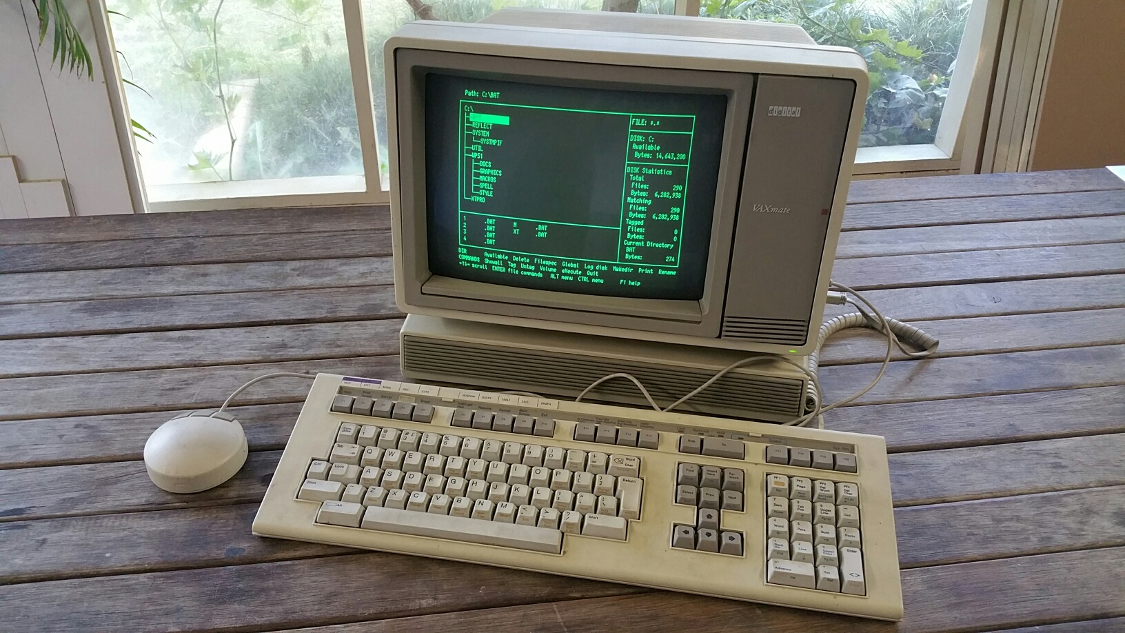 Grey computer with screen built in. A full-size keyboard in front and a round mouse on the side. The screen is showing green characters on a black background. Some file navigation?